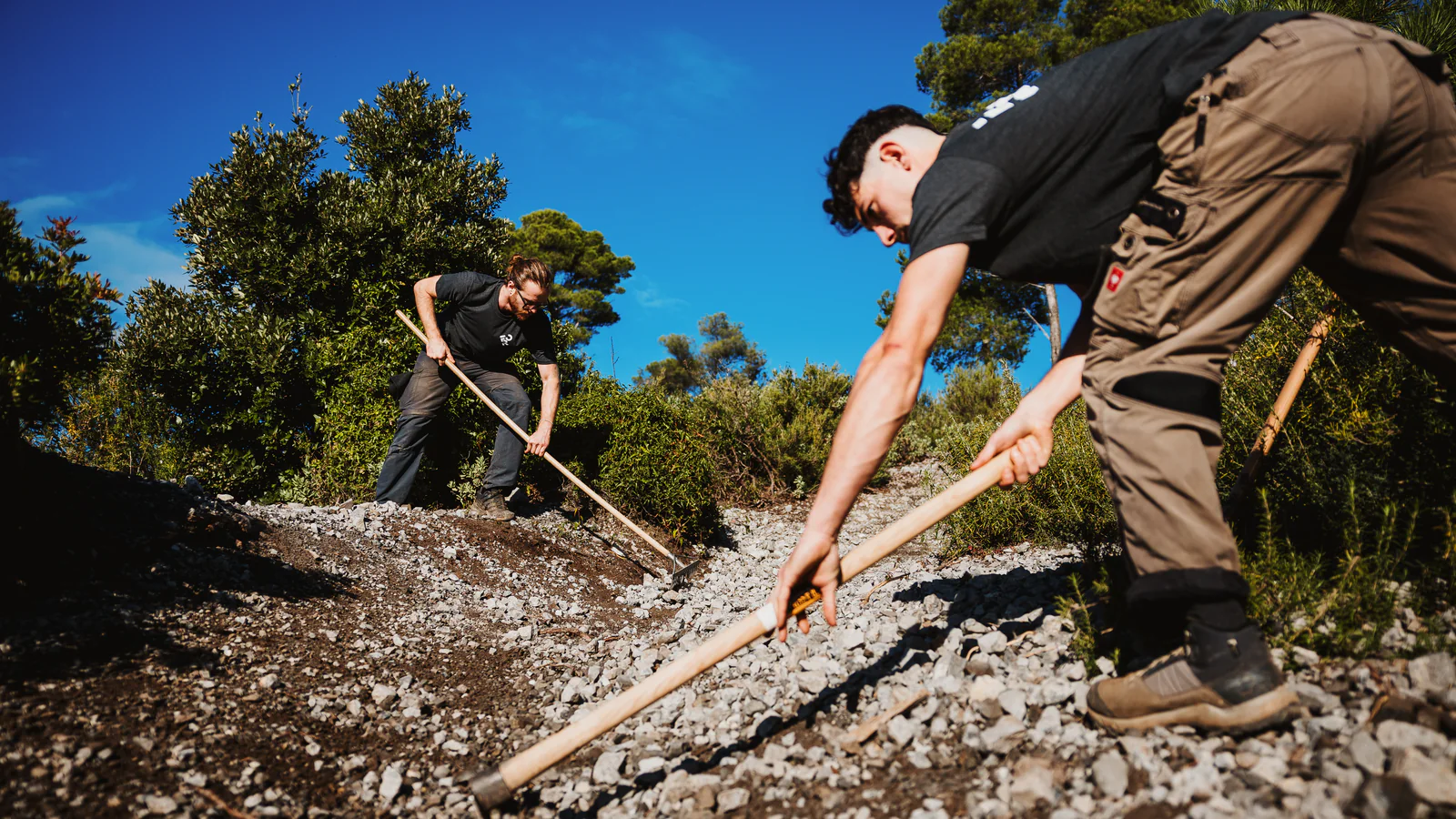 Two trail builders with rakes spread fresh gravel on a trail in the FINALE OUTDOOR REGION, under a blue sky